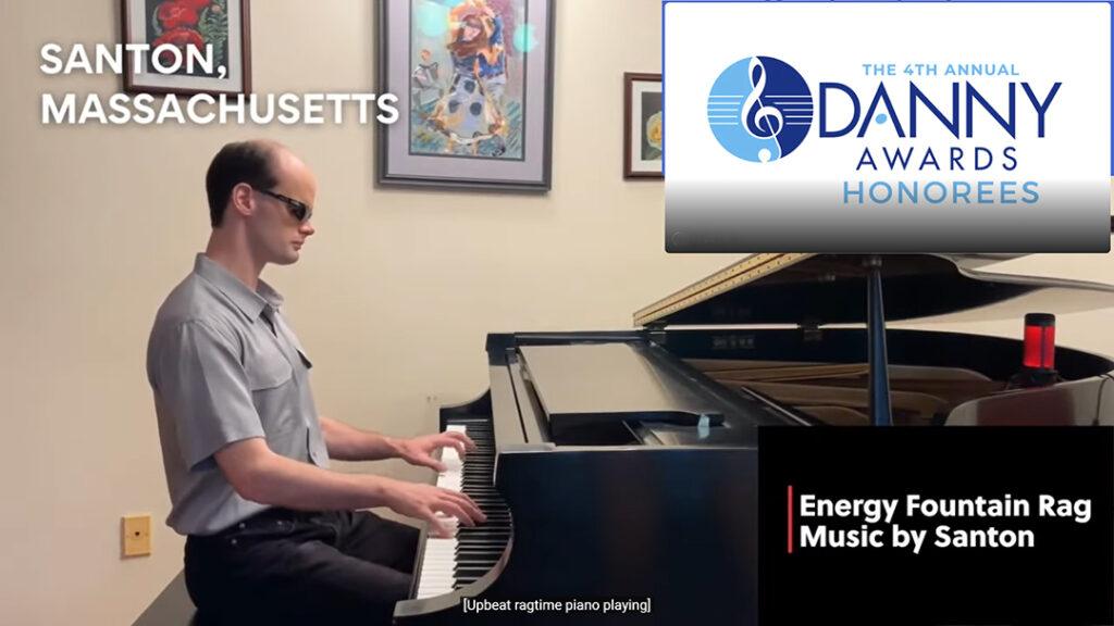 Santon dressed in a gray short-sleeved shirt, black pants and sunglasses plays grand piano at home. 
In the top right corner of the image, there is a treble clef as a DMF logo with the text: The 4th Annual Danny Awards Honorees". The text in the bottom right corner says "Energy Fountain Rag, music by Santon". In the top left corner of the image, the text says "Santon, Massachusetts".